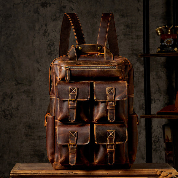 A vintage-style leather backpack with multiple front pockets, displayed against a dark textured background. The bag features brass accents and adjustable straps.