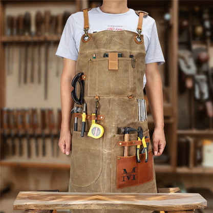 Person wearing khaki waxed canvas work apron with leather accents, tool pockets, workshop background
