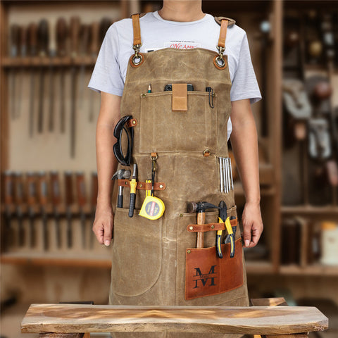 Person wearing khaki waxed canvas work apron with leather accents, tool pockets, workshop background