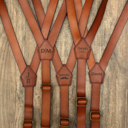 Multiple brown leather suspenders with personalized engravings (JR, DM, David, etc.), on wood background

