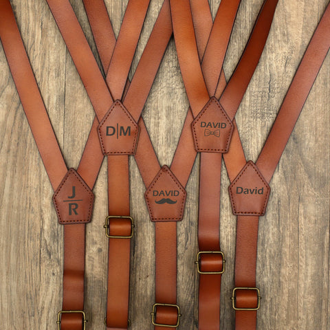 Multiple brown leather suspenders with personalized engravings (JR, DM, David, etc.), on wood background
