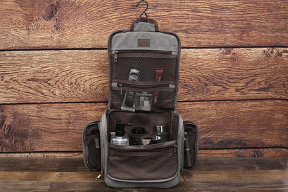 Grey canvas toiletry bag, fully open with hanging hook, multiple compartments filled with grooming products, wooden background