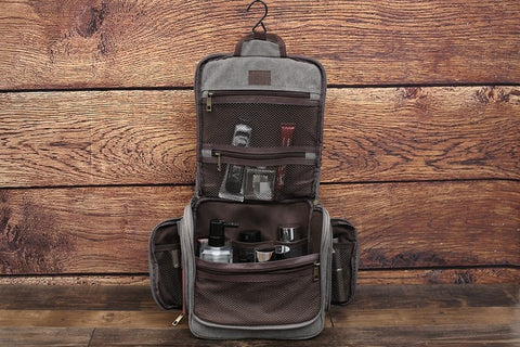Grey canvas toiletry bag, fully open with hanging hook, multiple compartments filled with grooming products, wooden background