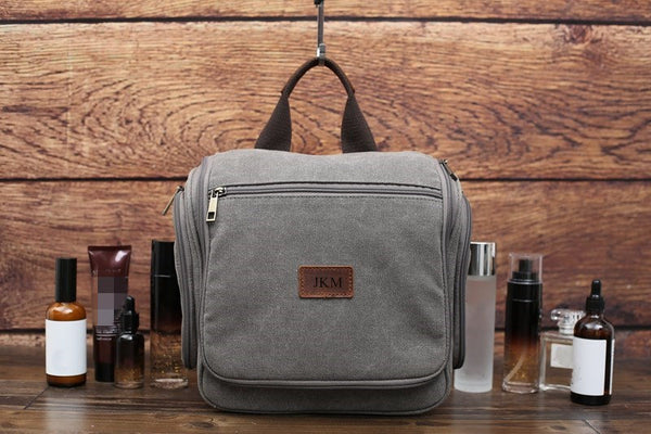Grey canvas toiletry bag, front view with "JKM" leather patch, surrounded by grooming products, wooden background