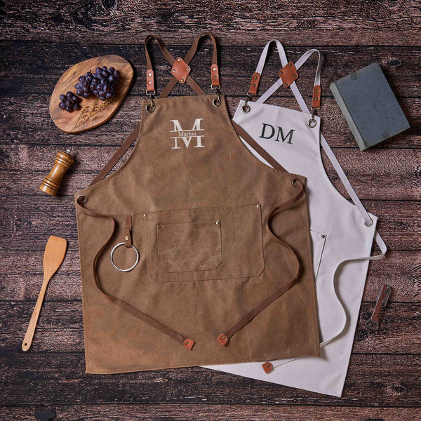 Two canvas aprons (brown with "Marlon", white with "DM"), flat lay with grapes, kitchen utensils, wooden background