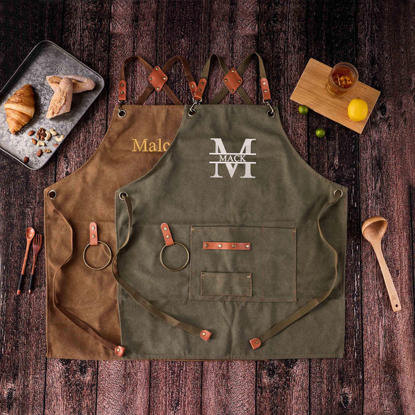 Two canvas aprons (brown with "Malc", olive with "MACK"), flat lay with kitchen food, wooden background
