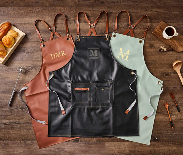 Three PU leather aprons (brown with "DMR", black with "M Morton", mint with "M Miller"), flat lay with pastries, coffee, kitchen utensils, wooden background