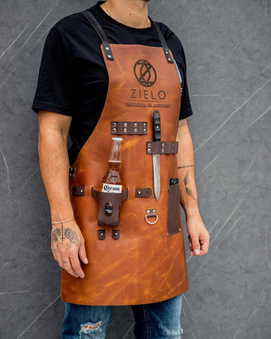 Man wearing brown leather apron with logo, holds Corona beer & knife, grey background
